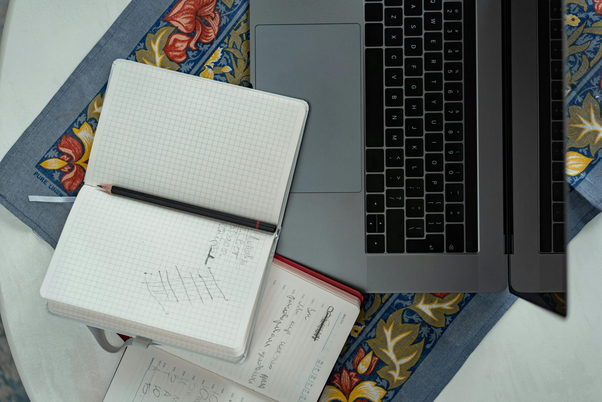 Flat lay of a laptop, notebooks, and a pencil on a floral tablecloth, ideal for productivity themes.
