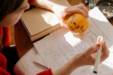 pexels-photo-4778676-4778676 Student studying math creatively using an orange and notes in warm indoor light.